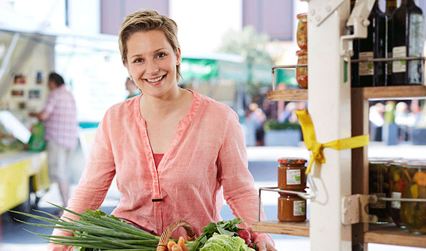Foto von Janine Steeger, die lächelnd an einem Tisch mit einem Einkaufskorb mit frischem Gemüse steht.