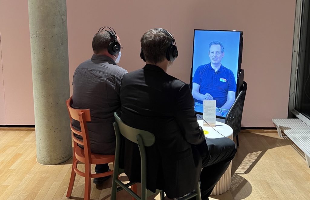 Eckart von Hirschhause auf einem Screen bei der Ausstellung Klima_X. Vor dem Screen sitzen zwei Besucher mit Kopfhörern
