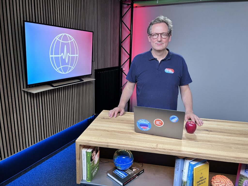 DGNB Jahreskongress 2023: Videostill der Keynote von Eckart von Hirschhausen, auf dem er hinter einem Holztisch mit Laptop und Apfel in eine Kamera blickt. Links neben ihm hängt ein Bildschirm mit einem Logo von GEGM. Er spricht über nachhaltiges Bauen.